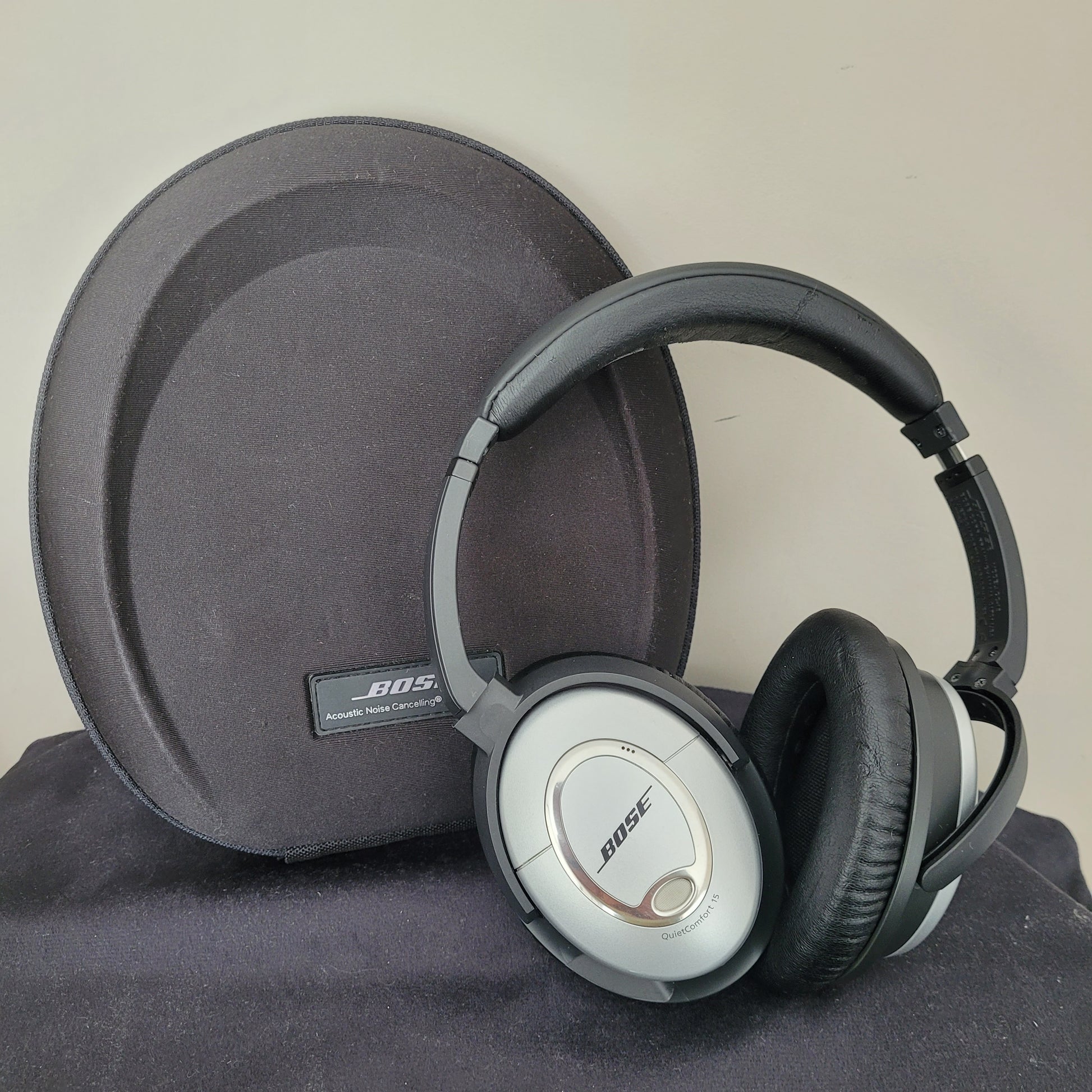 Die Bose Kopfhörer QuietComfort 15 liegen auf einer schwarzen, ovalen Tragetasche und präsentieren sich im klassischen Vintage-Design. Die silbernen Ohrmuscheln mit schwarzen Polstern unterstreichen den hochwertigen Look, während das Bose-Logo und die Modellbezeichnung „QuietComfort 15“ gut sichtbar sind. Die Tragetasche mit Bose-Branding vervollständigt das Set – ein Must-have für Liebhaber von Vintage-Elektronik und erstklassigen Kopfhörern.