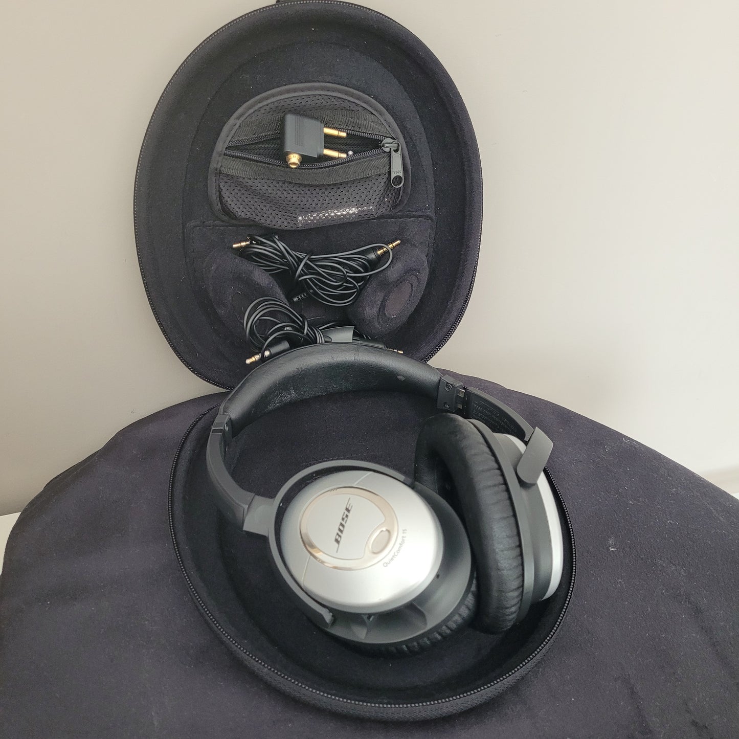 Das Bose Kopfhörer QuietComfort 15 Set ist geöffnet und zeigt das Zubehör innerhalb der schwarzen, ovalen Tragetasche. Der silberne Kopfhörer mit schwarzen Ohrpolstern liegt sicher in der Tasche, während das passende abnehmbare Audiokabel und der Airline-Adapter daneben platziert sind. Die hochwertige Verarbeitung und das durchdachte Zubehör unterstreichen die erstklassige Qualität dieses Vintage-Kopfhörers – ideal für Sammler und Liebhaber klassischer Elektronik.