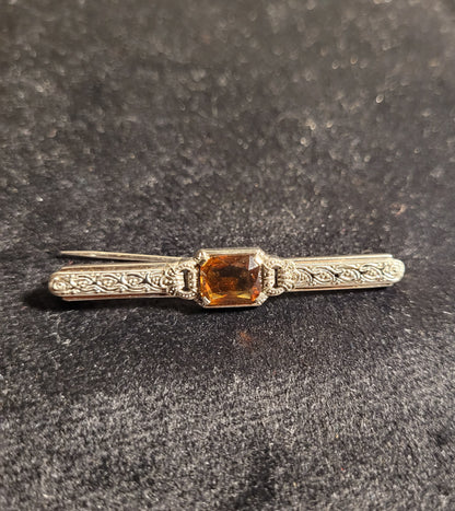 Antike silberne Brosche mit filigranen Verzierungen und rechteckigem, orangefarbenem Edelstein auf schwarzem Hintergrund.