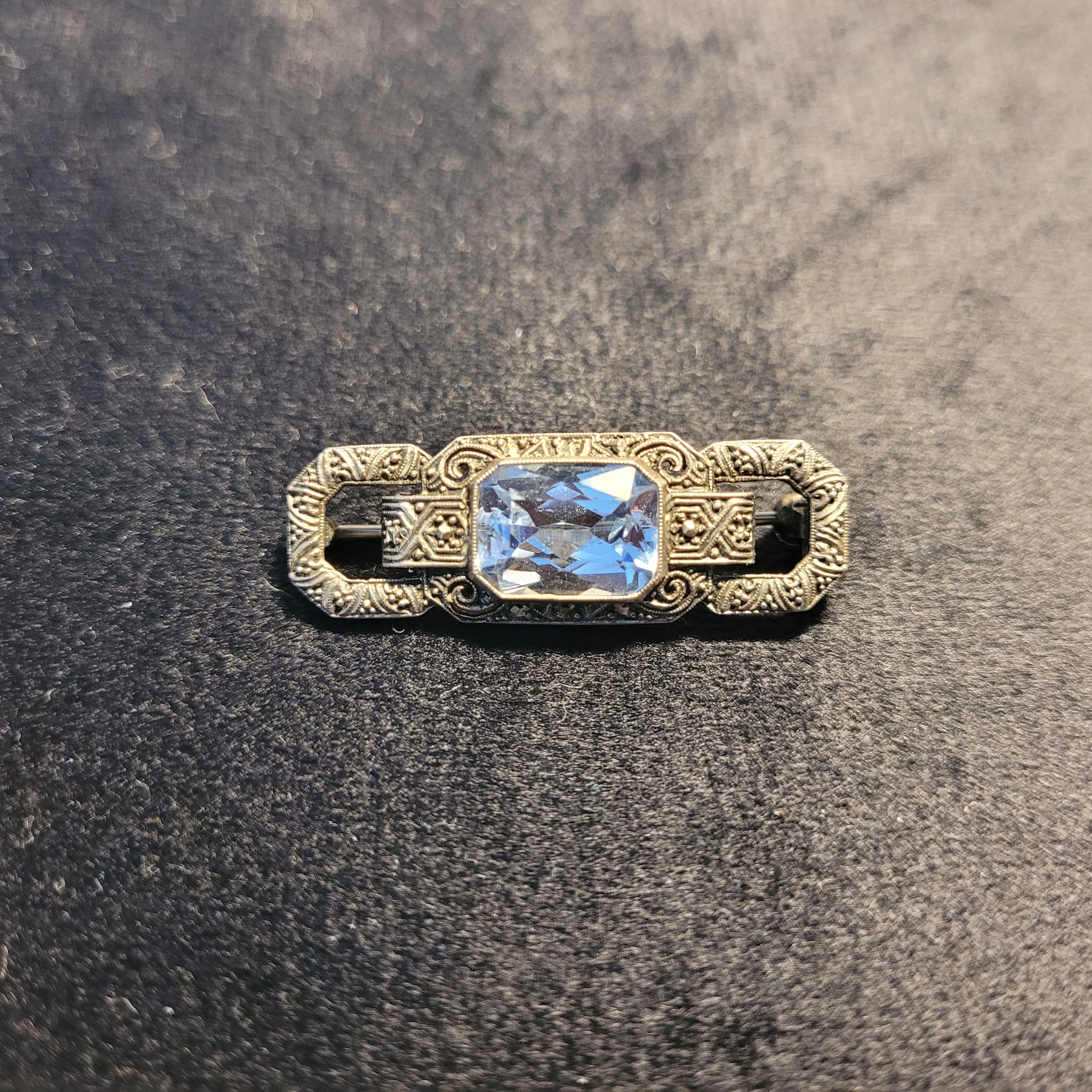 Vorderseite einer antiken Brosche mit rechteckigem, facettiertem blauem Edelstein und kunstvollem metallischen Design, verziert mit detaillierten Gravuren und Mustern