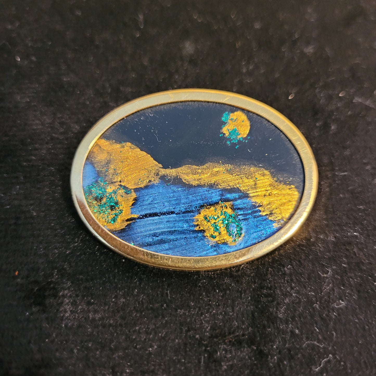 Ovale Brosche mit abstraktem Design in Blau- und Goldtönen, auf schwarzem Hintergrund. Das Kunstwerk zeigt leuchtende Farben und interessante Texturen, die an eine kosmische Landschaft erinnern.