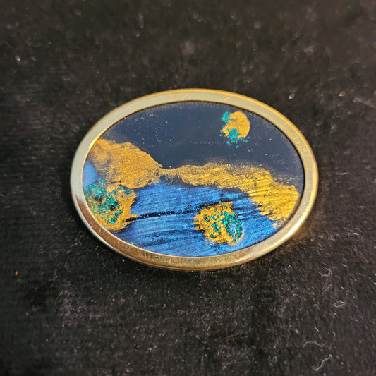 Ovale Brosche mit abstraktem Design in Blau- und Goldtönen, auf schwarzem Hintergrund. Das Kunstwerk zeigt leuchtende Farben und interessante Texturen, die an eine kosmische Landschaft erinnern.