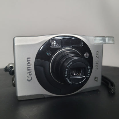 Silberne Canon IXUS Z70 Kompaktkamera mit ausgefahrenem Objektiv und integriertem Blitz, elegantes Design mit schwarzen Akzenten um das Objektiv.