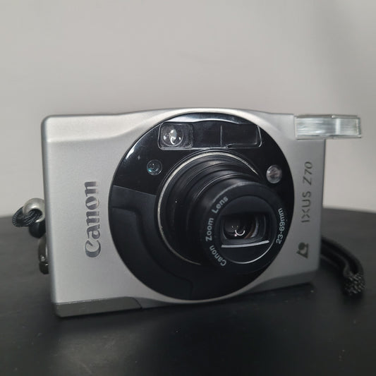Silberne Canon IXUS Z70 Kompaktkamera mit ausgefahrenem Objektiv und integriertem Blitz, elegantes Design mit schwarzen Akzenten um das Objektiv.