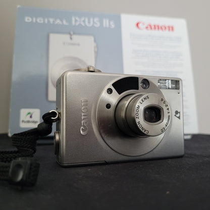 Nahaufnahme der silberfarbenen Canon Vintage Kamera Ixus II S mit Canon Zoom Lens 23–46mm, inklusive originaler Verpackung im Hintergrund – ideal für Sammler und Liebhaber klassischer Digitalkameras.