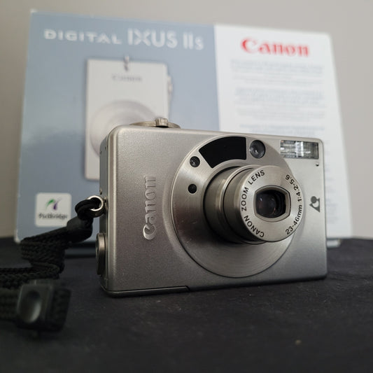 Nahaufnahme der silberfarbenen Canon Vintage Kamera Ixus II S mit Canon Zoom Lens 23–46mm, inklusive originaler Verpackung im Hintergrund – ideal für Sammler und Liebhaber klassischer Digitalkameras.