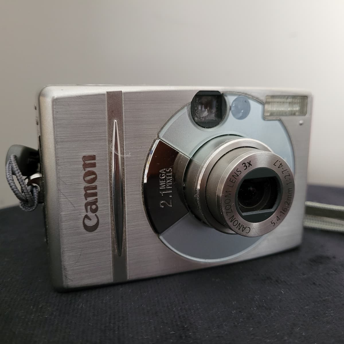 Canon IXY Digital 300 Digitalkamera in Silber, 3-fach Zoomobjektiv, 2,1 Megapixel, auf dunkler Oberfläche, mit Handschlaufe.