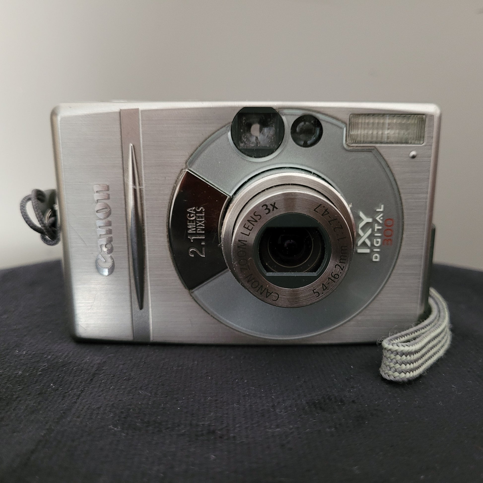 Canon IXY Digital 300 Digitalkamera in Silber, 3-fach Zoomobjektiv, 2,1 Megapixel, auf dunkler Oberfläche, mit Handschlaufe.