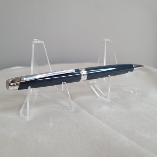Ein eleganter Caran d'Ache Druckbleistift Leman in Grau-Blau, präsentiert auf einem transparenten Ständer. Der Druckbleistift zeichnet sich durch stilvolles Design und silberne Akzente aus, was die schweizerische Präzision und exzellente Handwerkskunst betont. Ideal als luxuriöses Geschenk oder exklusives Accessoire für das Büro. Swiss Made.