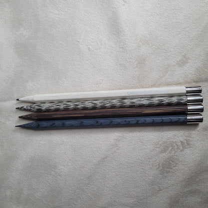 Set aus vier hochwertigen Caran d'Ache Bleistiften mit palladiumbeschichteten Kappen, präsentiert auf einer hellen, texturierten Oberfläche. Jeder Bleistift hat eine einzigartige Farbe und ein individuelles Muster. Von oben nach unten sind die Bleistifte: ein weißer Bleistift, ein weiß-schwarz gesprenkelter Bleistift, ein dunkelbrauner Bleistift mit Holzmaserung und ein dunkelblauer Bleistift mit Holzmaserung. Alle Bleistifte haben metallische silberne Kappen.