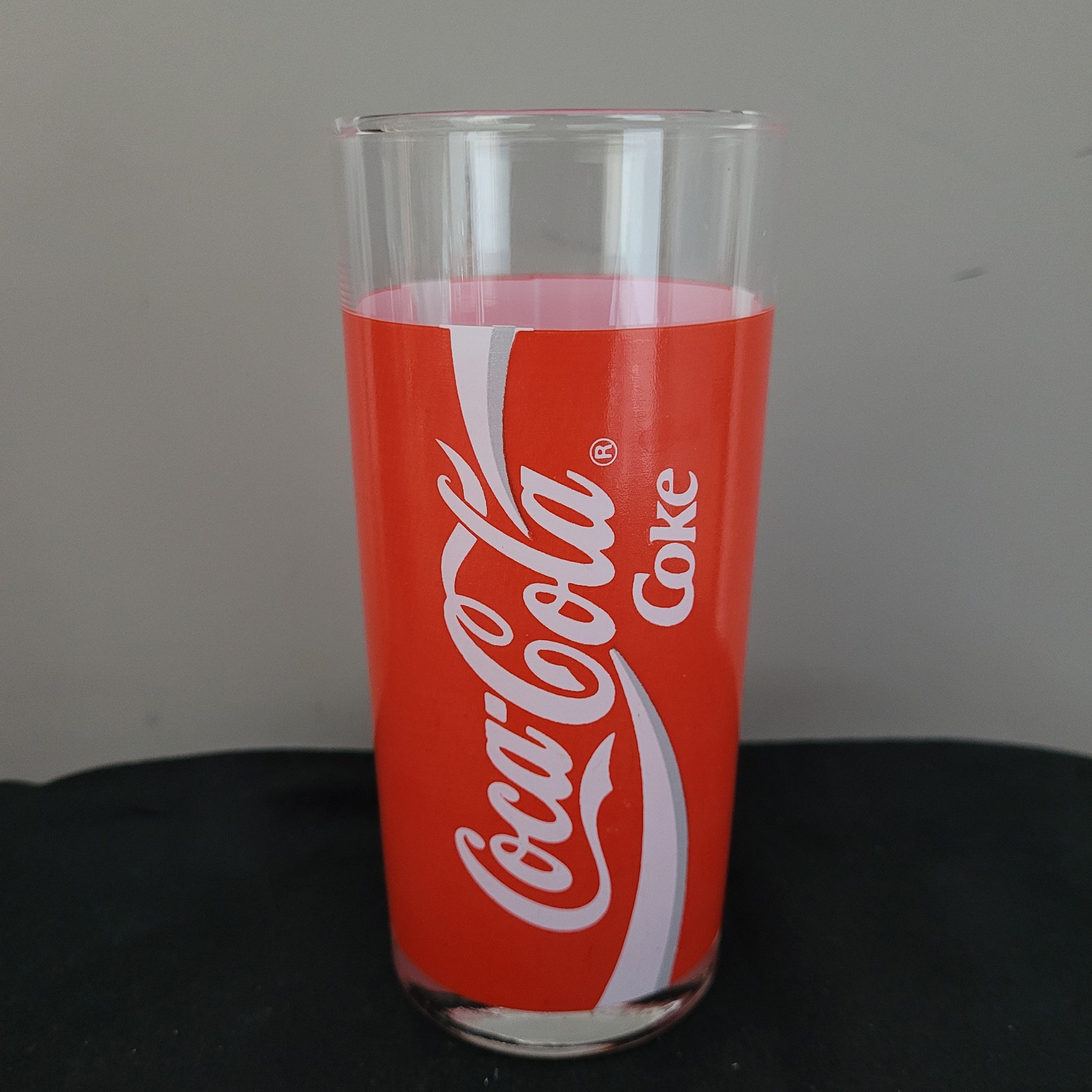 Ein einzelnes Coca-Cola Vintage Glas liegt seitlich auf einer schwarzen Oberfläche. Das Glas ist transparent und trägt eine rote Banderole mit dem weißen Schriftzug 'Coca-Cola' und 'Coke'. Der Hintergrund ist grau gehalten, was den Fokus auf das Glas und sein ikonisches Retro-Design lenkt.