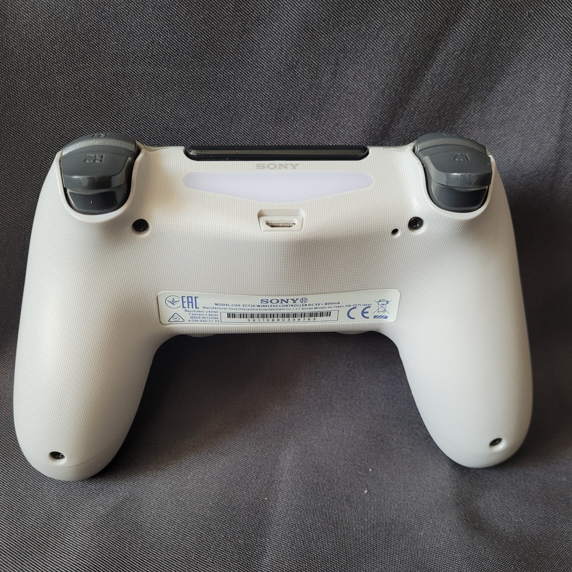 Rückseite eines Controller "Camouflage" – PlayStation 4 / PS4 – DualShock 4 – Original Sony in grau‑schwarz‑weißem Tarnmuster, mit sichtbaren L1‑ und R1‑Schultertasten, Lightbar, Micro‑USB‑Ladeanschluss und Modellaufkleber, liegend auf dunkler Stoffoberfläche. EAN 0711719453512 / Modell CUH-ZCT1E