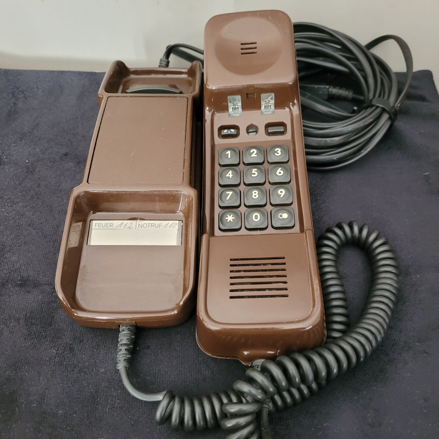 Braunes Vintage-Dallas -Telefon mit Wähltasten, spiralförmigem Kabel und Notrufhinweisen auf schwarzem Stoff.