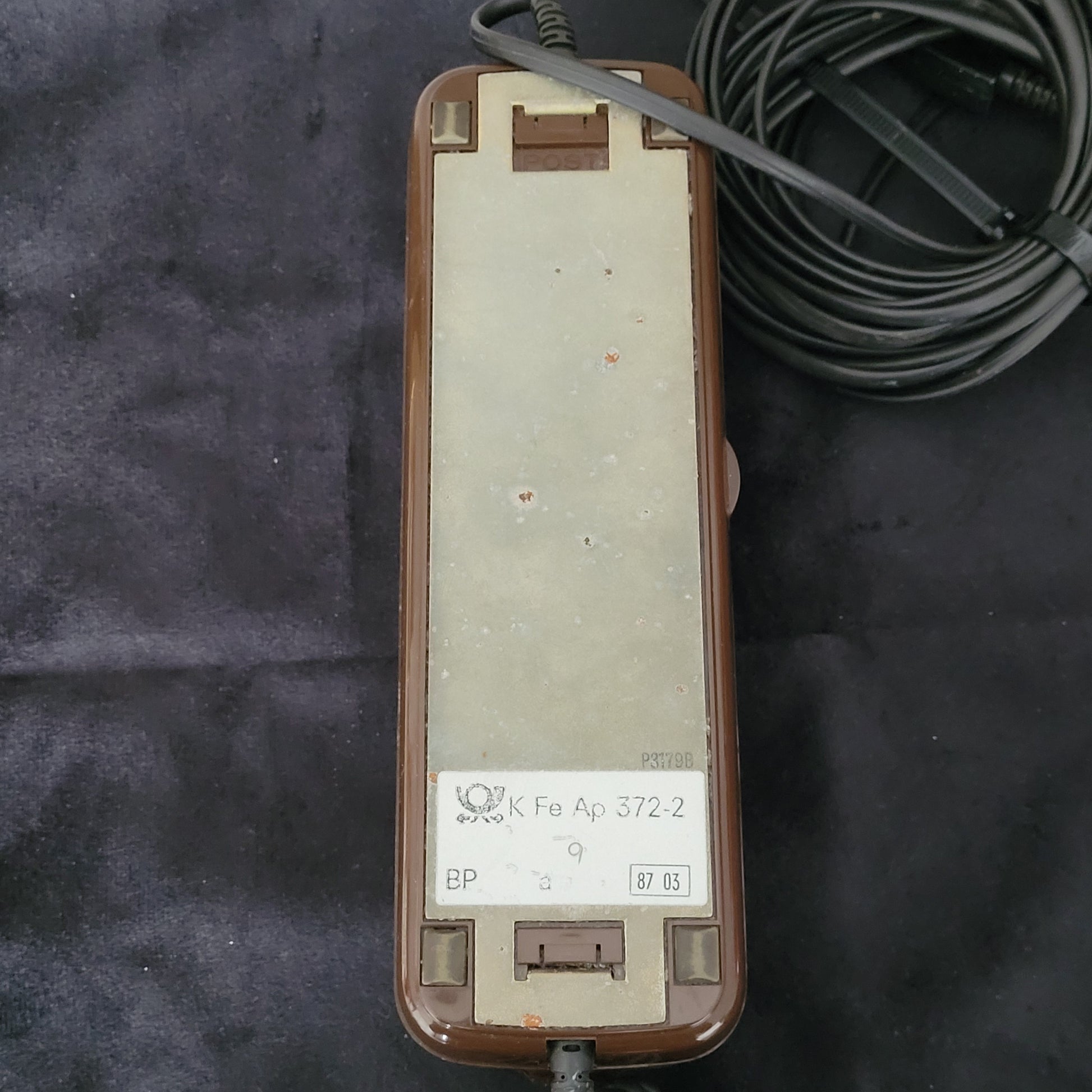 Unterseite eines braunen Vintage-Telefons mit Metallschild und spiralförmigem Kabel.