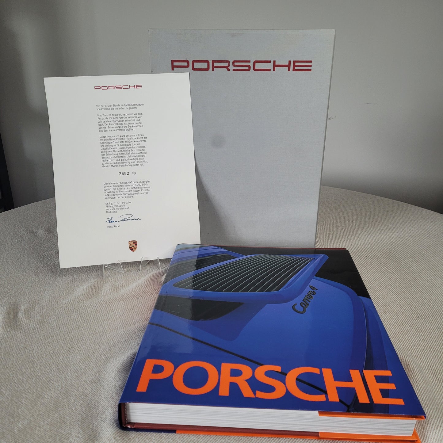 Arrangement von Porsche-Materialien: Aufgeschlagenes Buch mit blauem Cover und orangefarbenem "PORSCHE"-Schriftzug, Porsche-Brief und graue Mappe mit rotem Porsche-Logo im Hintergrund
