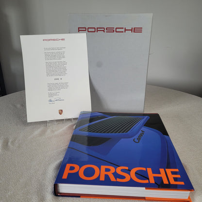 Arrangement von Porsche-Materialien: Aufgeschlagenes Buch mit blauem Cover und orangefarbenem "PORSCHE"-Schriftzug, Porsche-Brief und graue Mappe mit rotem Porsche-Logo im Hintergrund