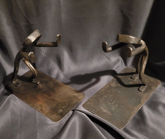 Paar handgeschmiedete Vintage Buchstützen von Höller im brutalistischen Design. Die massiven Eisenstützen zeigen zwei abstrahierte, kraftvolle Figuren in stützender Pose auf einer flachen Metallbasis, perfekt für schwere Bücher.
