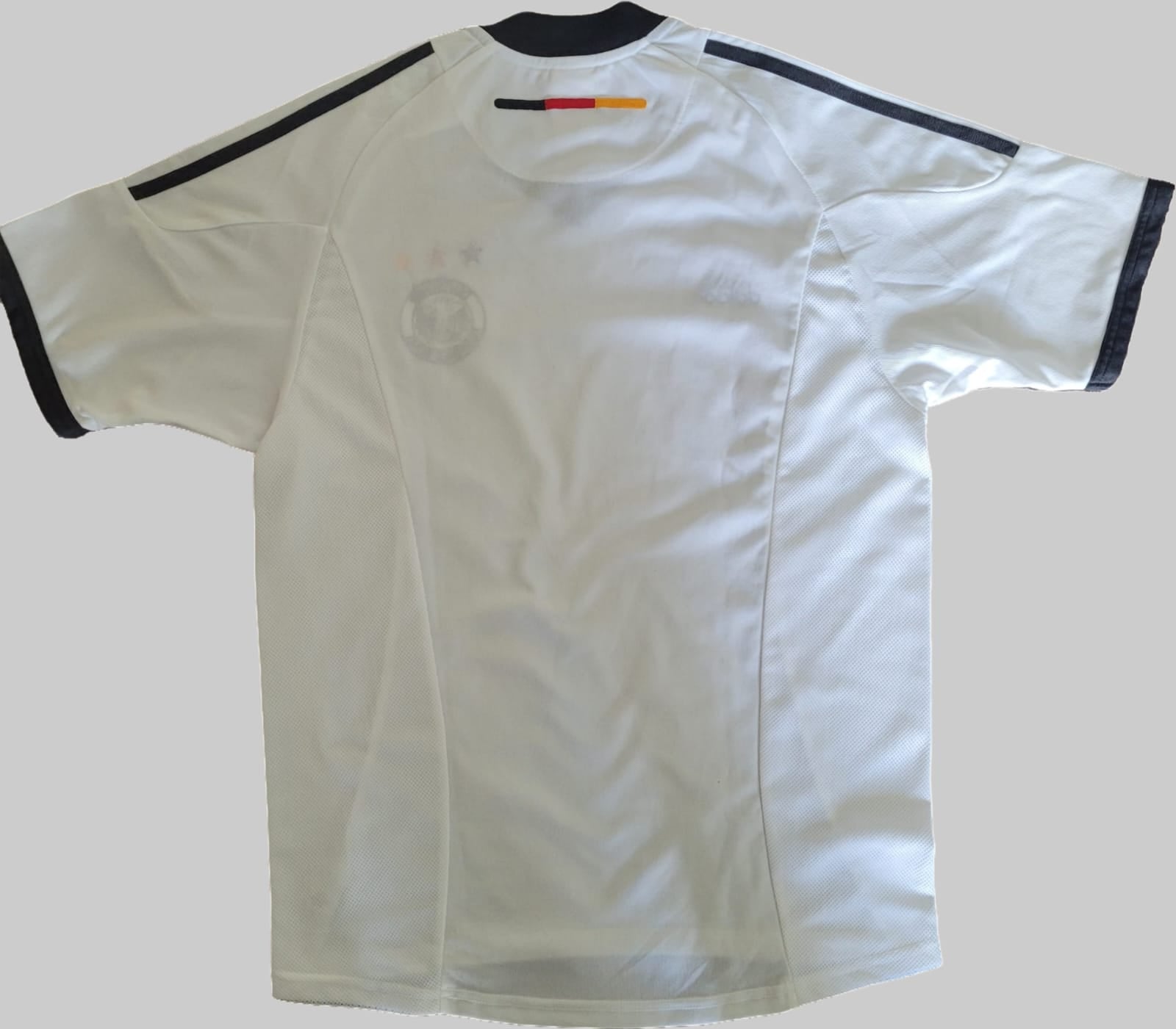 Deutschland Vintage Trikot 2002 – Rückseite auf grauem Hintergrund Das Bild zeigt die Rückseite eines Deutschland Vintage Trikots aus der WM 2002, platziert auf einem grauen Hintergrund. Das Trikot ist weiß mit schwarzen Akzenten an Kragen und Ärmeln. Die Rückseite ist schlicht gehalten, ohne sichtbare Rückennummer oder Schriftzüge