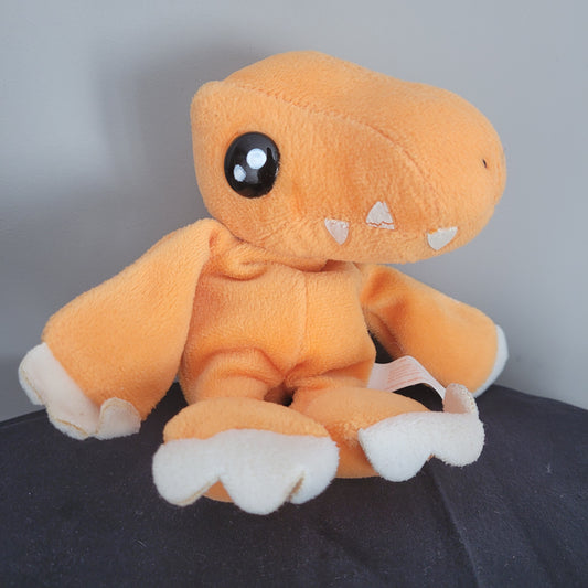 Ein orangefarbenes Plüschtier von Digimon, das wie ein Dinosaurier Agumon aussieht, liegt auf einer dunklen Oberfläche. Das Plüschtier hat große schwarze Augen und weiße Zähne, die aus seinem Maul ragen. Es hat auch weiße Krallen an den Pfoten.