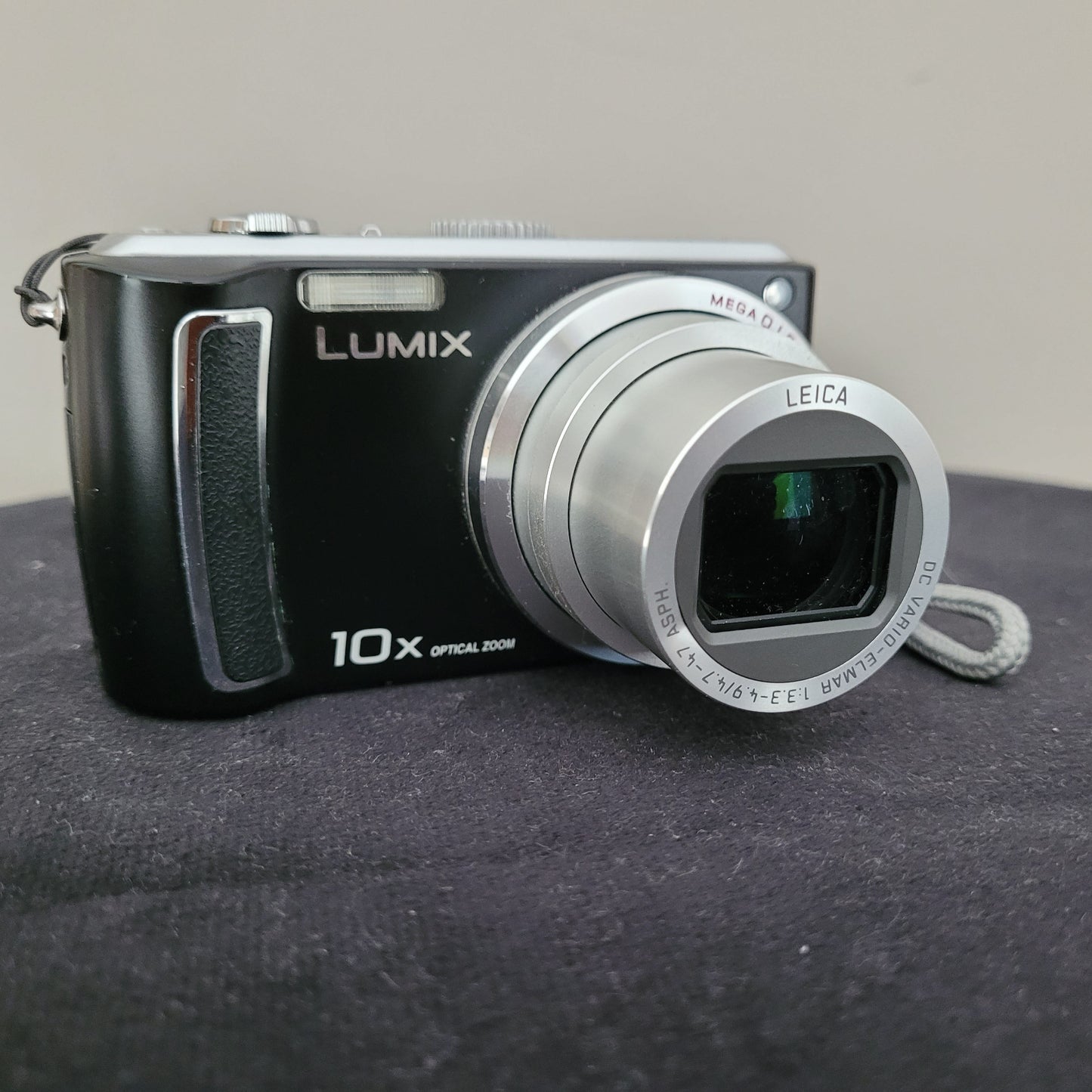 Die Panasonic Lumix DMC-TZ5 Digitalkamera liegt auf einem schwarzen Untergrund mit weißem Hintergrund. Auf der Kamera sind verschiedene Beschriftungen erkennbar, darunter „MEGA O.I.S.“, „LEICA“, „DC VARIO-ELMAR“, „10x OPTICAL ZOOM“ und „LUMIX“. Das Objektiv der Kamera ist in ausgefahrenem Zustand.