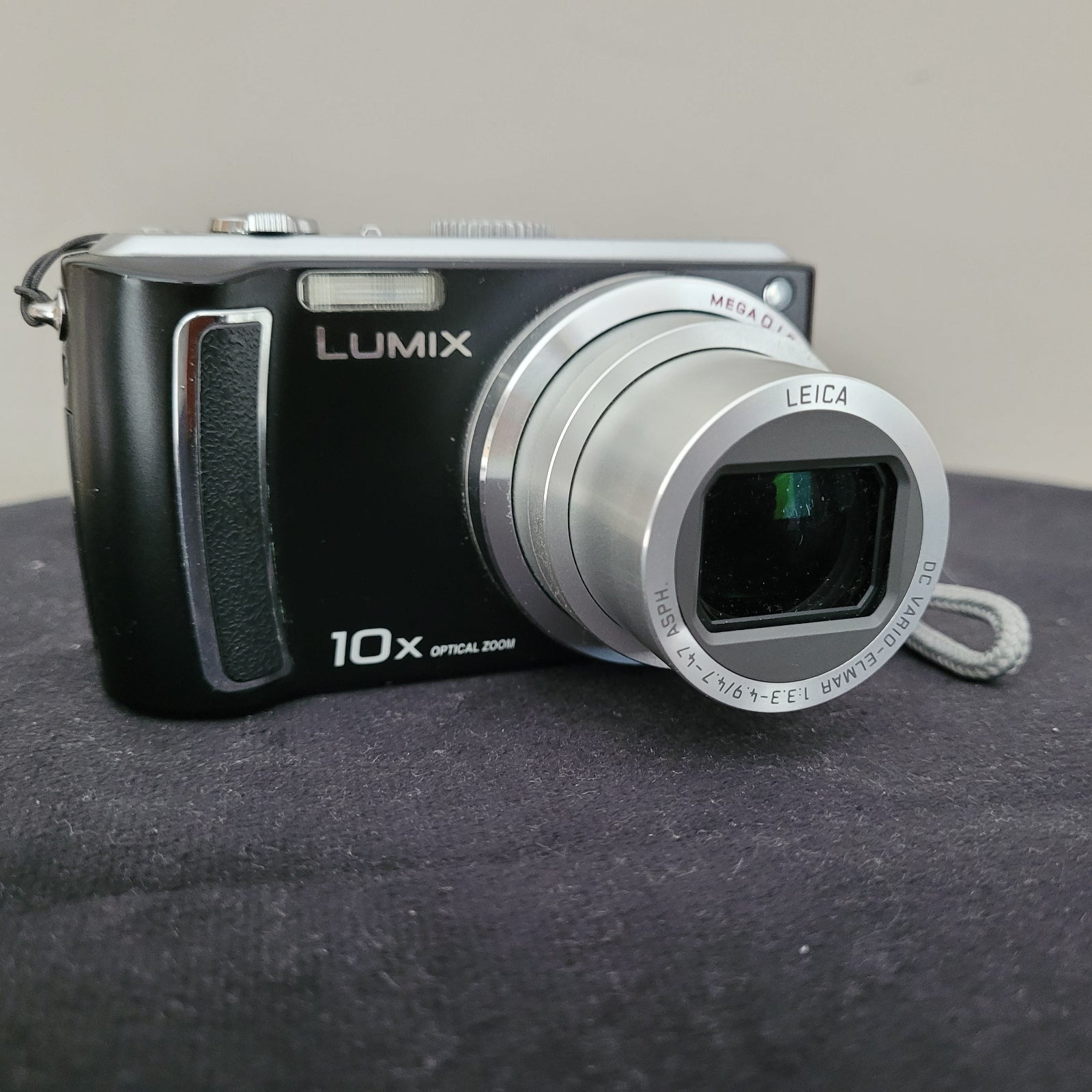 Die Panasonic Lumix DMC-TZ5 Digitalkamera liegt auf einem schwarzen Untergrund mit weißem Hintergrund. Auf der Kamera sind verschiedene Beschriftungen erkennbar, darunter „MEGA O.I.S.“, „LEICA“, „DC VARIO-ELMAR“, „10x OPTICAL ZOOM“ und „LUMIX“. Das Objektiv der Kamera ist in ausgefahrenem Zustand.
