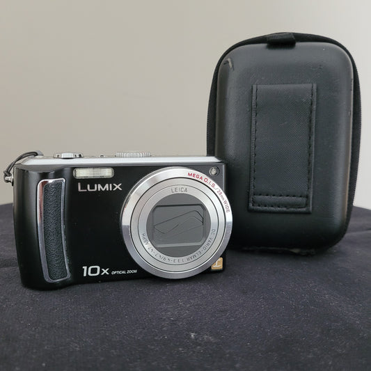 Die Panasonic Lumix DMC-TZ5 Digitalkamera liegt auf einem schwarzen Untergrund mit weißem Hintergrund. Auf der Kamera sind verschiedene Beschriftungen erkennbar, darunter „MEGA O.I.S.“, „LEICA“, „DC VARIO-ELMAR“, „10x OPTICAL ZOOM“ und „LUMIX“. Daneben befindet sich eine schwarze Kameratasche.