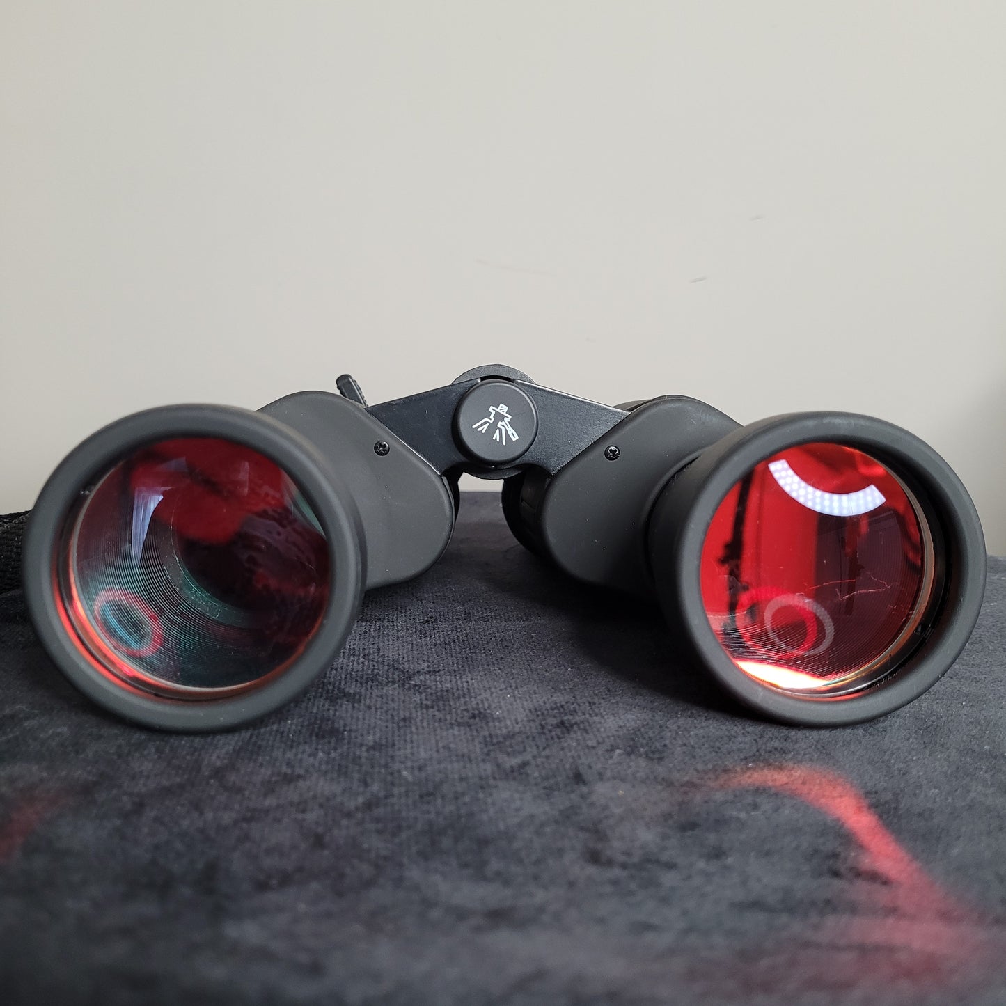 Ein Prismatic 8-24x50 Zoom Fernglas mit auffälligen roten Linsen, das auf einer dunklen Oberfläche liegt. Das Fernglas hat ein schwarzes Gehäuse und ein kleines Symbol, das wie ein Vogel oder eine Feder aussieht, ist auf dem Scharnier zu sehen. Die roten Linsen reflektieren Licht und zeigen kreisförmige Muster.