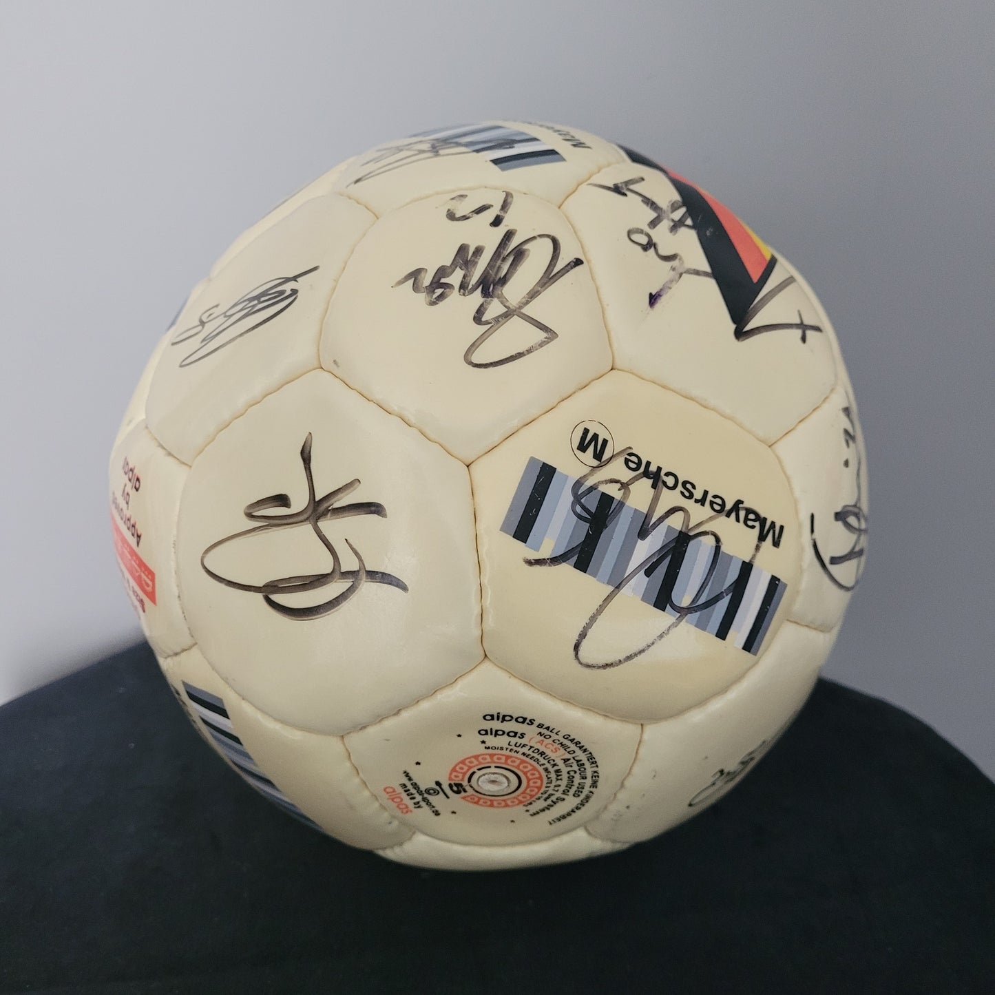 TSV Alemannia Aachen Vintage Fußball – Seitenansicht auf schwarzer Oberfläche Das Bild zeigt die Seitenansicht eines gelben Fußballs mit Original-Unterschriften, platziert auf einer schwarzen Oberfläche mit grauem Hintergrund. Die Balloberfläche weist sichtbare Gebrauchsspuren auf, die den Vintage-Charakter des Sammlerstücks unterstreichen. Die Unterschriften sind gut erkennbar und verleihen dem Fußball besonderen Sammlerwert.