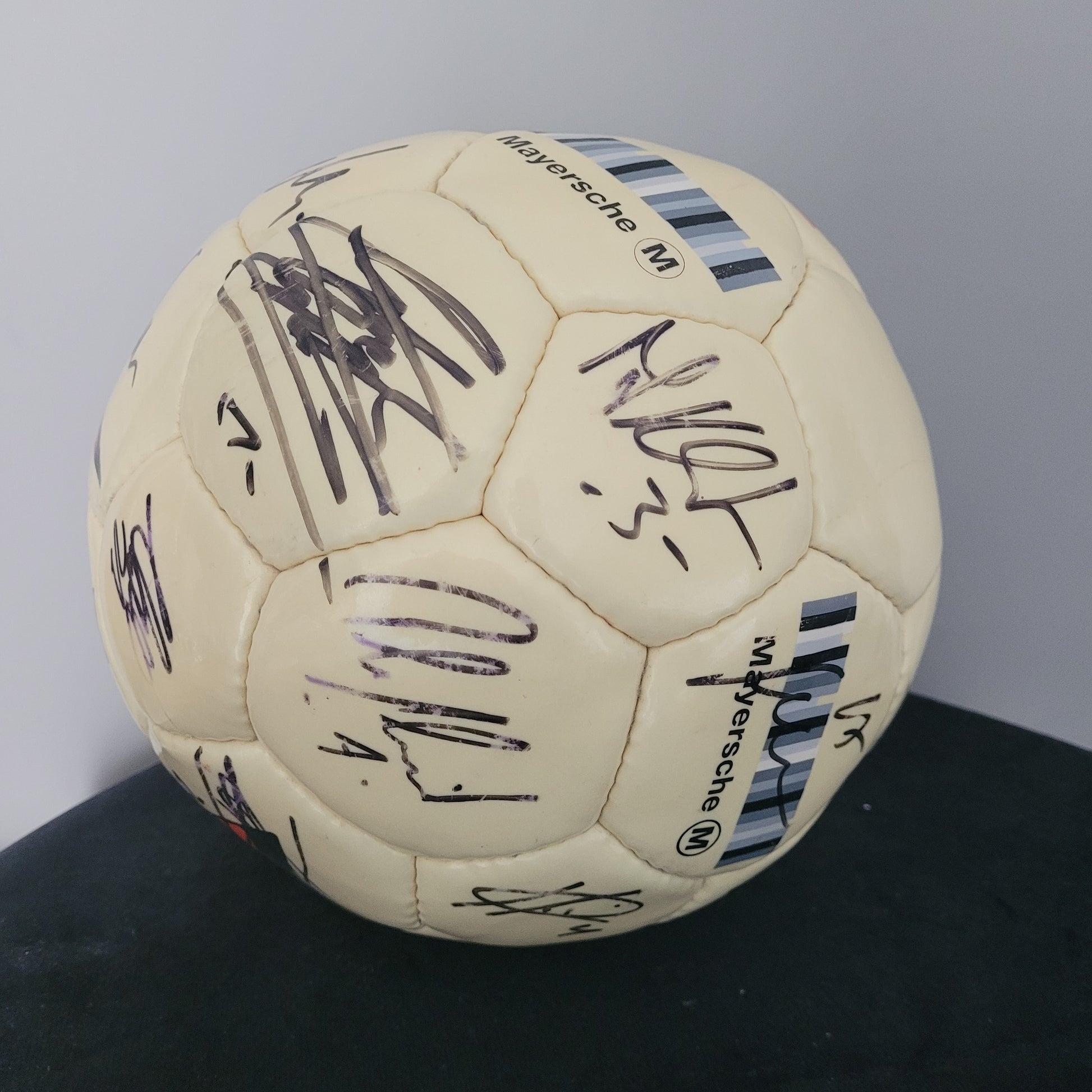 TSV Alemannia Aachen Vintage Fußball – Seitenansicht auf schwarzer Oberfläche Das Bild zeigt die Seitenansicht eines gelben Fußballs mit Original-Unterschriften, platziert auf einer schwarzen Oberfläche mit grauem Hintergrund. Die Balloberfläche weist sichtbare Gebrauchsspuren auf, die den Vintage-Charakter des Sammlerstücks unterstreichen. Die Unterschriften sind gut erkennbar und verleihen dem Fußball besonderen Sammlerwert.