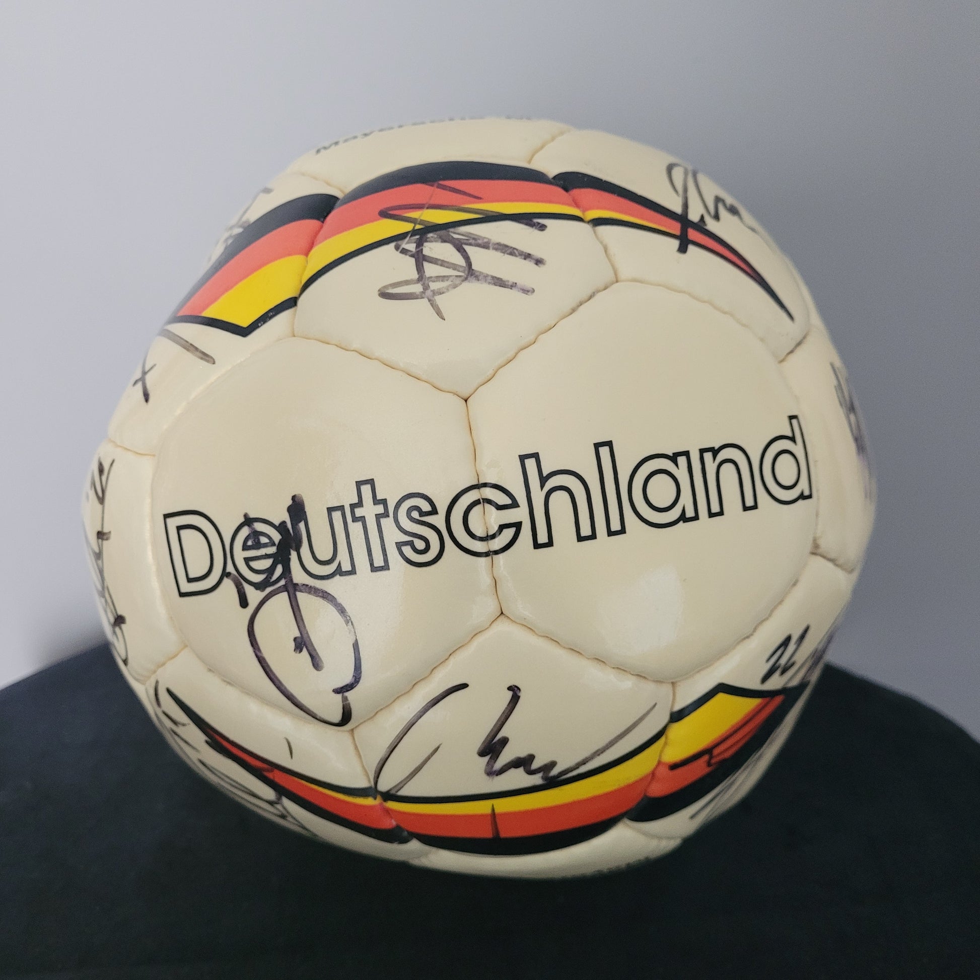 TSV Alemannia Aachen Vintage Fußball – auf schwarzer Oberfläche mit grauem Hintergrund Das Bild zeigt einen Fußball mit einer gelben Grundfarbe, auf einer schwarzen Oberfläche mit grauem Hintergrund platziert. Auf dem Ball sind mehrere Original-Unterschriften sichtbar, die sich deutlich vom Material abheben. Die Oberfläche weist einige sichtbare Gebrauchsspuren auf, die den Vintage-Charakter dieses Sammlerstücks unterstreichen.