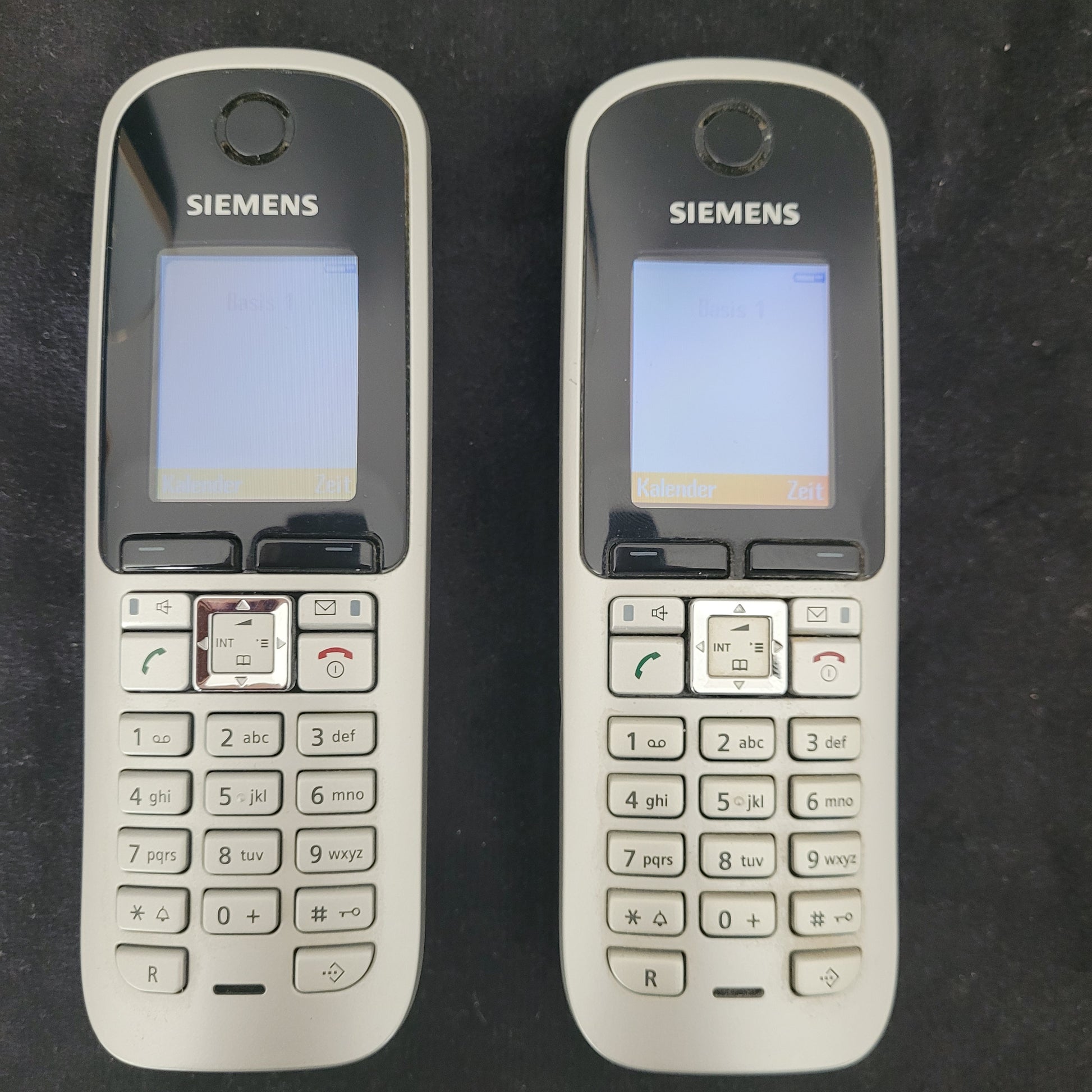 Die Gigaset Siemens S670 Duo Eco Dect Titanium Intuitive Eleganz Schnurlostelefone liegen nebeneinander auf einem dunklen Hintergrund. Die Vorderseite der Mobilteile zeigt das stilvolle Titanium-Finish, das klare LCD-Display und die intuitiven Bedienelemente. Das Haupttelefon ist eingeschaltet, und die Tasten sind gut sichtbar – eine elegante und moderne DECT-Telefonlösung für eine zuverlässige Kommunikation.
