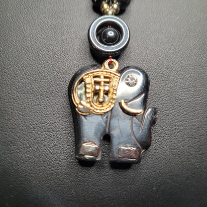 Anhänger in Form eines Elefanten aus glänzendem dunklem Material mit goldenen Verzierungen, an einer Kette befestigt, auf schwarzem Hintergrund.