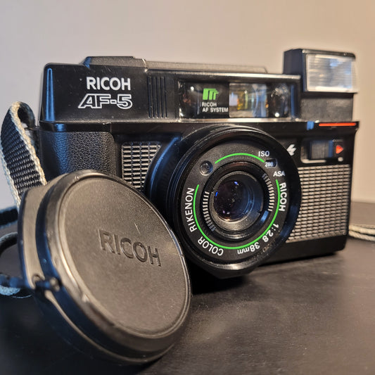 Nahaufnahme der Ricoh AF-5 Kamera mit Ricoh AF System und Rikenon 38mm f/2.8 Objektiv, inklusive Objektivdeckel im Vordergrund. Ideal für Fotografie-Enthusiasten.