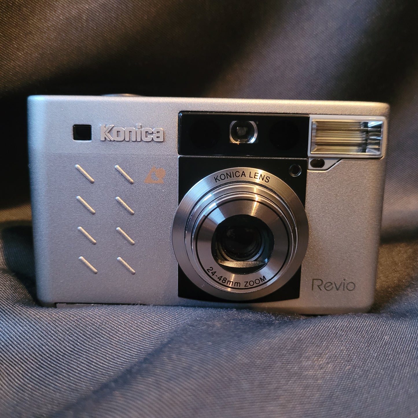 Vorderseite der Konica Revio Digitalkamera in silbernem Gehäuse mit Zoomobjektiv 2.4–48mm, Konica-Logo, Blitz, Sucher und Modellschriftzug. Vintage Kamera mit kompaktem Design.