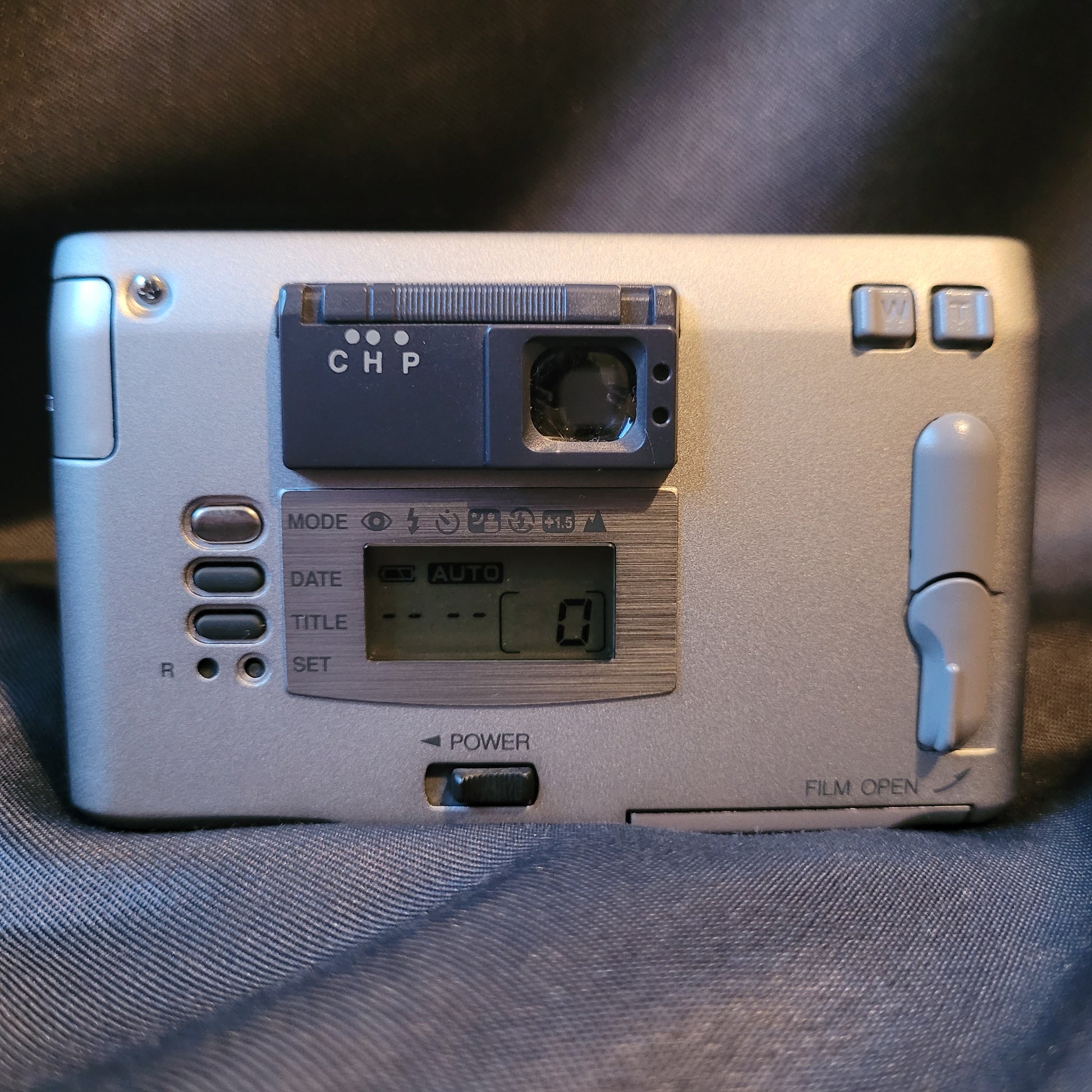 Rückseite der Konica Revio Kamera mit LCD-Display, Zoomtasten (W/T), Moduswahl, Datumsanzeige und Filmfachhebel. Vintage Analogkamera mit silbernem Gehäuse und Bedienelementen für APS-Film.