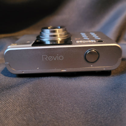 Obere Seite der Konica Revio Kamera mit ausgefahrenem Objektiv, silbernem Gehäuse und sichtbarem Auslöser. Vintage Digitalkamera mit kompaktem Design und Markenlogo.