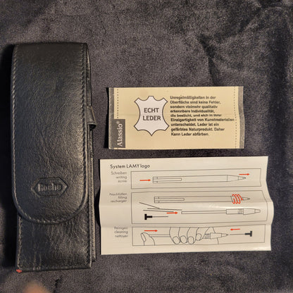 Ein schwarzes Lederetui mit einer geprägten Logoaufschrift liegt auf einem dunklen Stoffhintergrund. Daneben befindet sich eine beige Karte mit dem Text 'ECHT LEDER' und eine Anleitung zur Verwendung des Systems LAMY Logo.