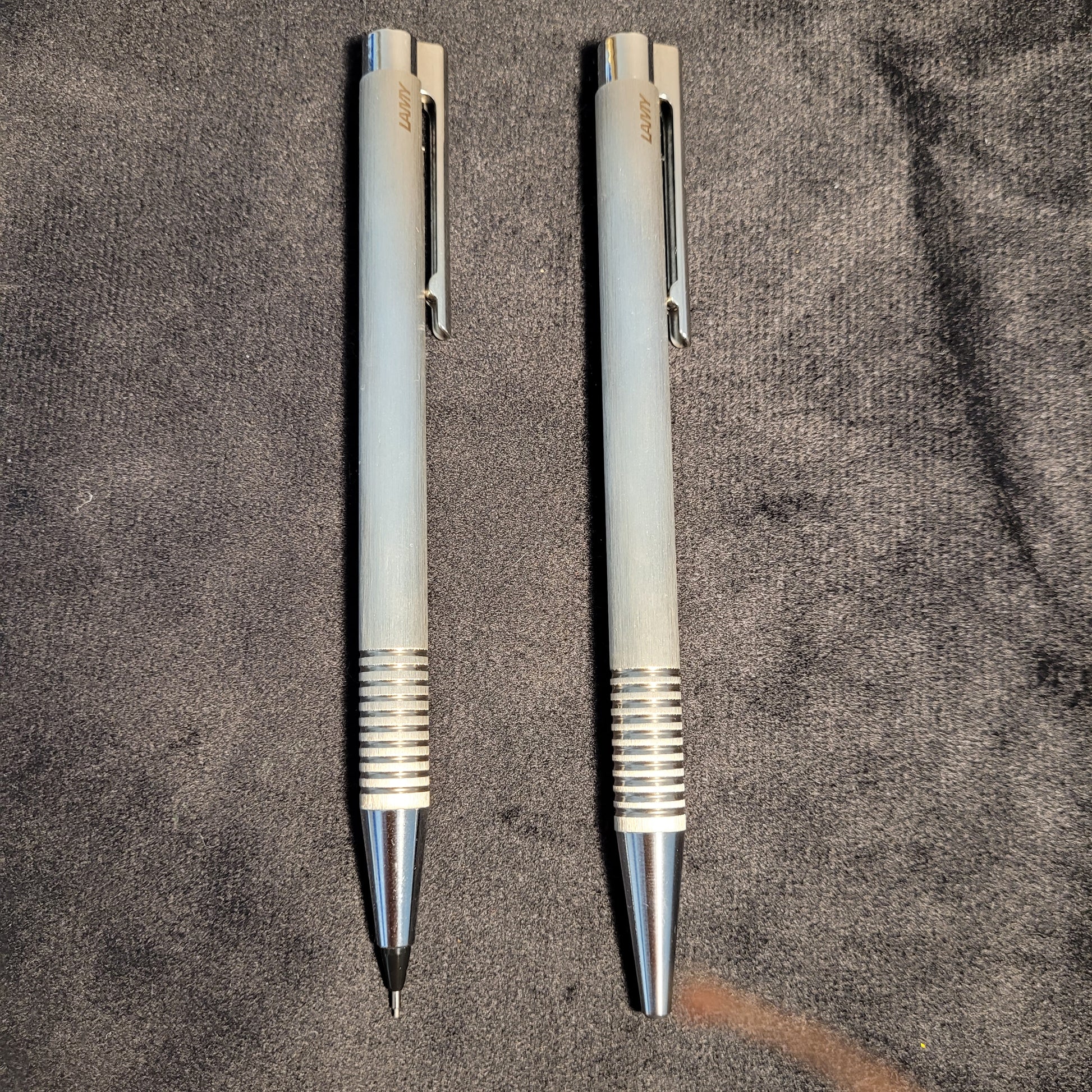 Zwei silberfarbene mechanische Stifte der Marke LAMY liegen parallel auf einem dunklen Stoffhintergrund. Beide Stifte haben eine geriffelte Griffzone und einen Clip mit dem eingravierten Markennamen. Einer der Stifte ist bereit zur Nutzung.