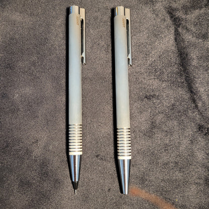 Zwei silberfarbene mechanische Stifte der Marke LAMY liegen parallel auf einem dunklen Stoffhintergrund. Beide Stifte haben eine geriffelte Griffzone und einen Clip mit dem eingravierten Markennamen. Einer der Stifte ist bereit zur Nutzung.