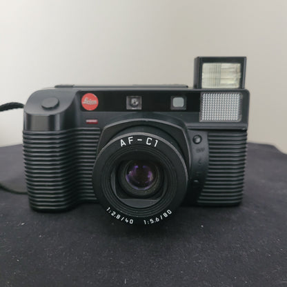Leica Vintage Analogkamera AF-C1 in Schwarz mit integriertem Blitz und Dual-Objektiv (40 mm f/2.8 & 80 mm f/5.6), präsentiert auf dunkler Oberfläche – klassische Leica Kompaktkamera für analoge Fotografie mit ikonischem Retro-Design und rotem Leica-Logo.
