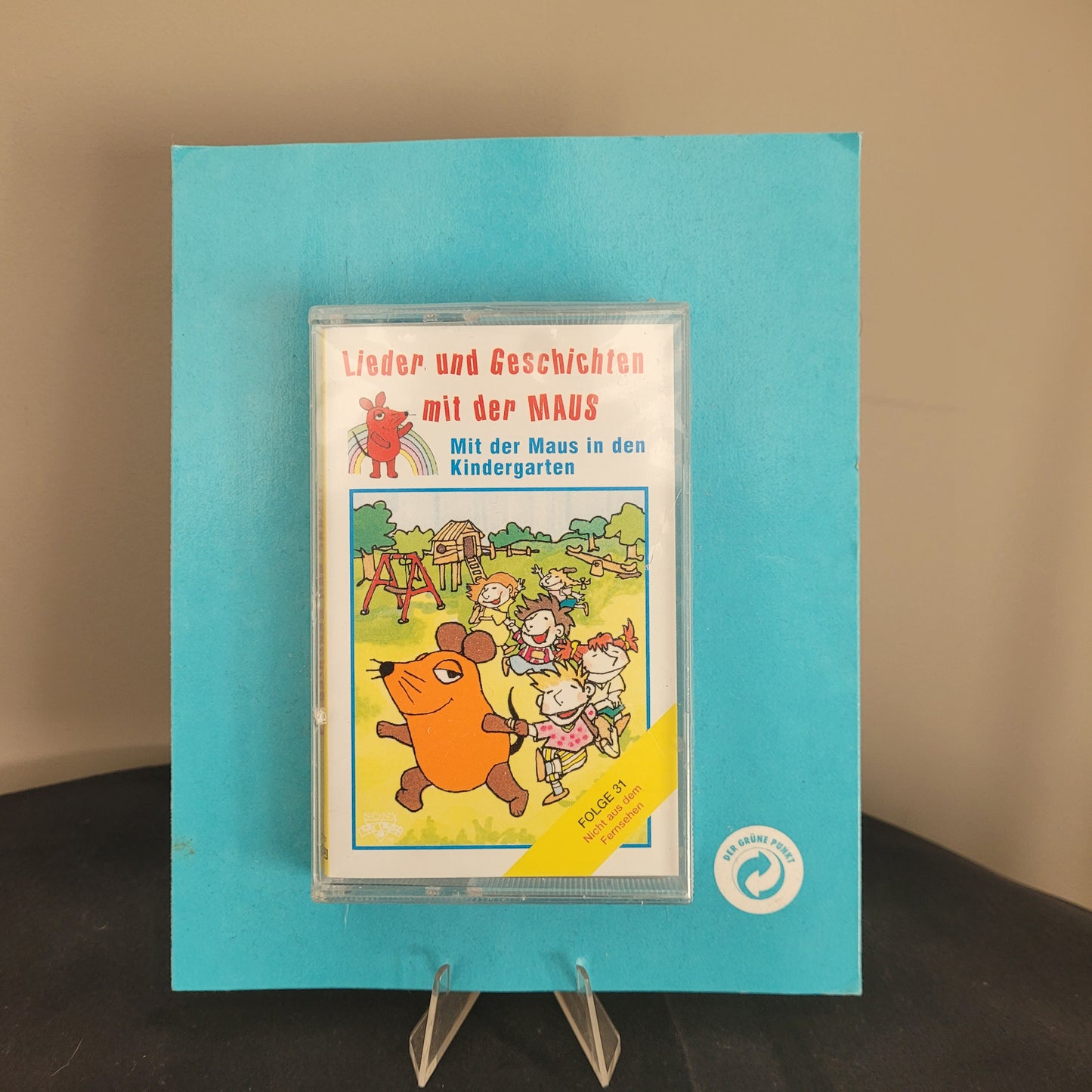 Das Bild zeigt eine Kassette mit dem Titel 'Lieder und Geschichten mit der MAUS - Mit der Maus in den Kindergarten'. Die Kassette befindet sich in einer durchsichtigen Hülle und liegt auf einem blauen Hintergrund. Auf dem Cover der Kassette ist eine Illustration von Kindern und der Maus in einem Kindergarten zu sehen. Diese Kassette ist ein nostalgisches Produkt, das sich an Kinder und Eltern richtet, die Geschichten und Lieder mit der beliebten Figur 'Die Maus' genießen möchten.