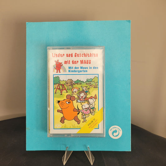 Das Bild zeigt eine Kassette mit dem Titel 'Lieder und Geschichten mit der MAUS - Mit der Maus in den Kindergarten'. Die Kassette befindet sich in einer durchsichtigen Hülle und liegt auf einem blauen Hintergrund. Auf dem Cover der Kassette ist eine Illustration von Kindern und der Maus in einem Kindergarten zu sehen. Diese Kassette ist ein nostalgisches Produkt, das sich an Kinder und Eltern richtet, die Geschichten und Lieder mit der beliebten Figur 'Die Maus' genießen möchten.