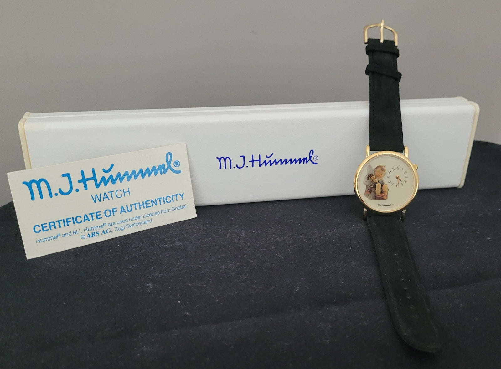 Das Bild zeigt die Vorderseite einer M.J. Hummel Armbanduhr mit einem eleganten, goldfarbenen Gehäuse und einem schwarzen Lederarmband. Im Zentrum stechen das kindlich illustrierte Zifferblatt und das daneben platzierte Zertifikat der Authentizität hervor, auf dem unter anderem die Unterschriften „M.J. Hummel WATCH CERTIFICATE OF AUTHENTICITY“ und Lizenzinformationen stehen. Im Hintergrund befindet sich eine weiße Box mit dem blauen M.J. Hummel Logo.
