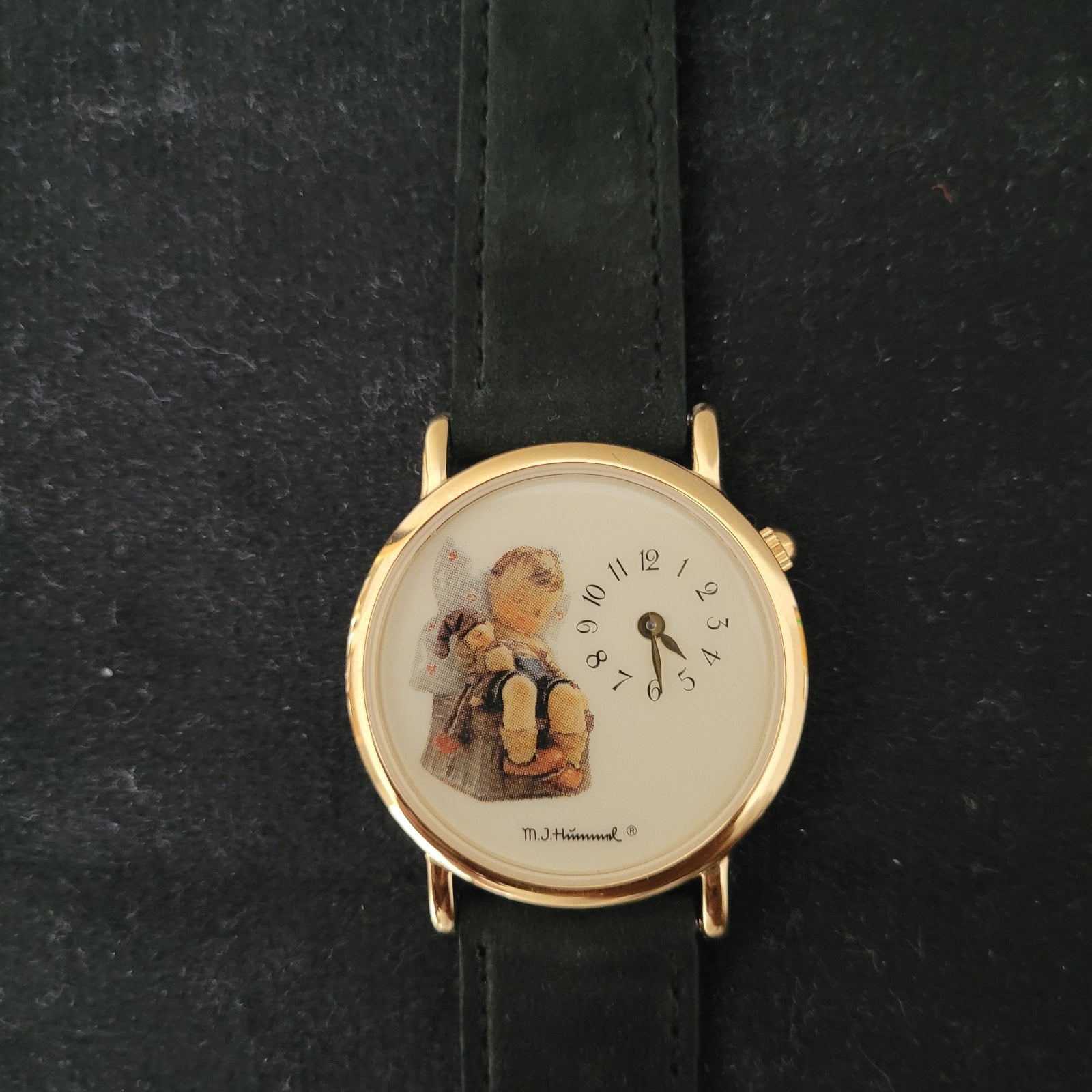Das Bild zeigt die Vorderseite einer M.J. Hummel Vintage Armbanduhr mit einem eleganten, goldfarbenen Gehäuse und einem schwarzen Lederarmband. Das kunstvoll gestaltete Zifferblatt ist mit einer Illustration eines Kindes, das eine Puppe hält, versehen, und rechts am Zifferblatt ist der Schriftzug „M.J. Hummel“ erkennbar – ein authentisches Original-Vintage-Design.