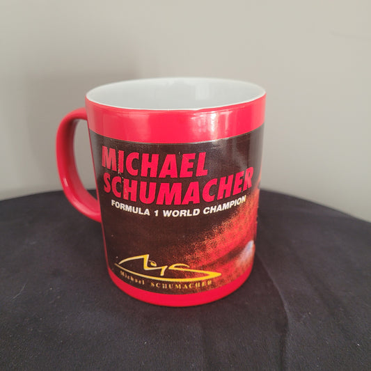 Eine rote Tasse mit dem Aufdruck 'MICHAEL SCHUMACHER' in großen roten Buchstaben auf schwarzem Hintergrund. Darunter steht 'FORMULA 1 WORLD CHAMPION' in kleineren weißen Buchstaben. Auf der rechten Seite der Tasse befindet sich eine stilisierte Unterschrift von Michael Schumacher in gelber Farbe. Die Tasse ist auf einem schwarzen Untergrund platziert.