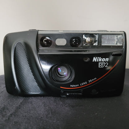 Eine hochwertige Nikon RF2 Kamera mit einem 35mm Objektiv, präsentiert mit einem eleganten schwarzen Gehäuse und einem markanten Griff. Neben der Kamera liegt eine stilvolle schwarze Nikon Kameratasche. Das Set liegt auf einer dunklen Oberfläche und der neutrale Hintergrund lenkt nicht vom Produkt ab.