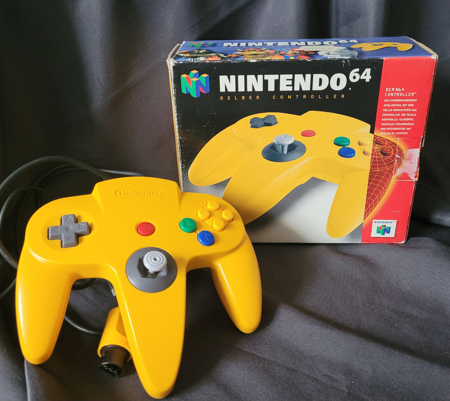 Nintendo 64 – N64 – Gelber Controller/Joystick Model NUS-005A mit Originalverpackung (OVP). Der Controller zeigt das klassische dreizackige Design mit grauem Analogstick, rotem Startknopf, Steuerkreuz sowie bunten A-, B- und C-Tasten. Die OVP trägt das Nintendo 64 Logo und die Aufschrift „Gelber Controller“ mit Abbildung des Controllers. Retro/Vintage Zubehör für die N64 Konsole.