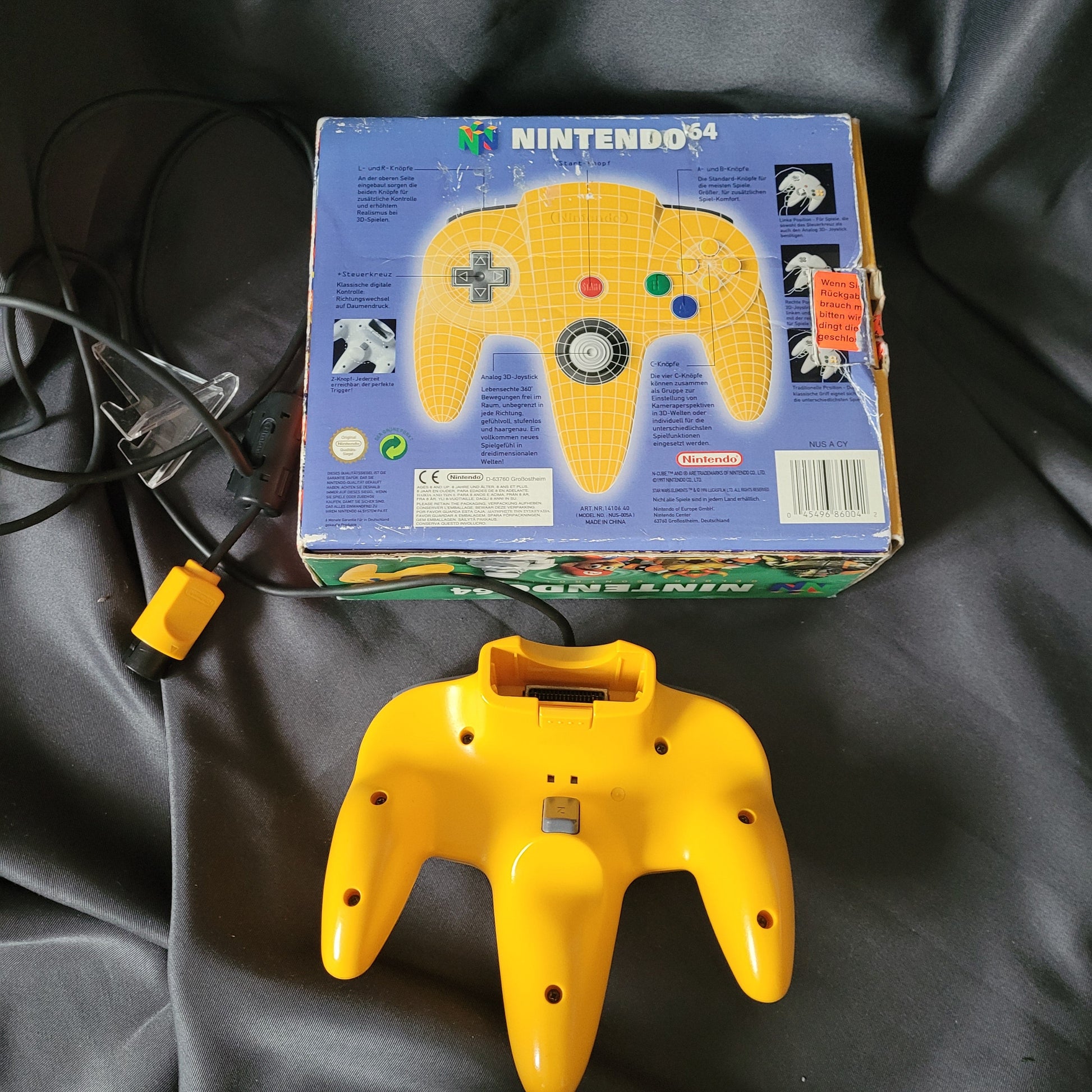 Nintendo 64 – N64 – Gelber Controller/Joystick Model NUS-005A mit Originalverpackung (OVP). Der kabelgebundene Controller mit dreizackigem Design, grauem Analogstick, rotem Startknopf, Steuerkreuz sowie bunten A-, B- und C-Tasten liegt vor der Rückseite der OVP. Auf der Verpackung sind das Nintendo 64 Logo, Produktinformationen in mehreren Sprachen sowie eine schematische Darstellung des Controllers mit Beschriftung der Tasten zu sehen. Retro/Vintage Zubehör für die N64 Konsole.