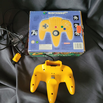 Nintendo 64 – N64 – Gelber Controller/Joystick Model NUS-005A mit Originalverpackung (OVP). Der kabelgebundene Controller mit dreizackigem Design, grauem Analogstick, rotem Startknopf, Steuerkreuz sowie bunten A-, B- und C-Tasten liegt vor der Rückseite der OVP. Auf der Verpackung sind das Nintendo 64 Logo, Produktinformationen in mehreren Sprachen sowie eine schematische Darstellung des Controllers mit Beschriftung der Tasten zu sehen. Retro/Vintage Zubehör für die N64 Konsole.