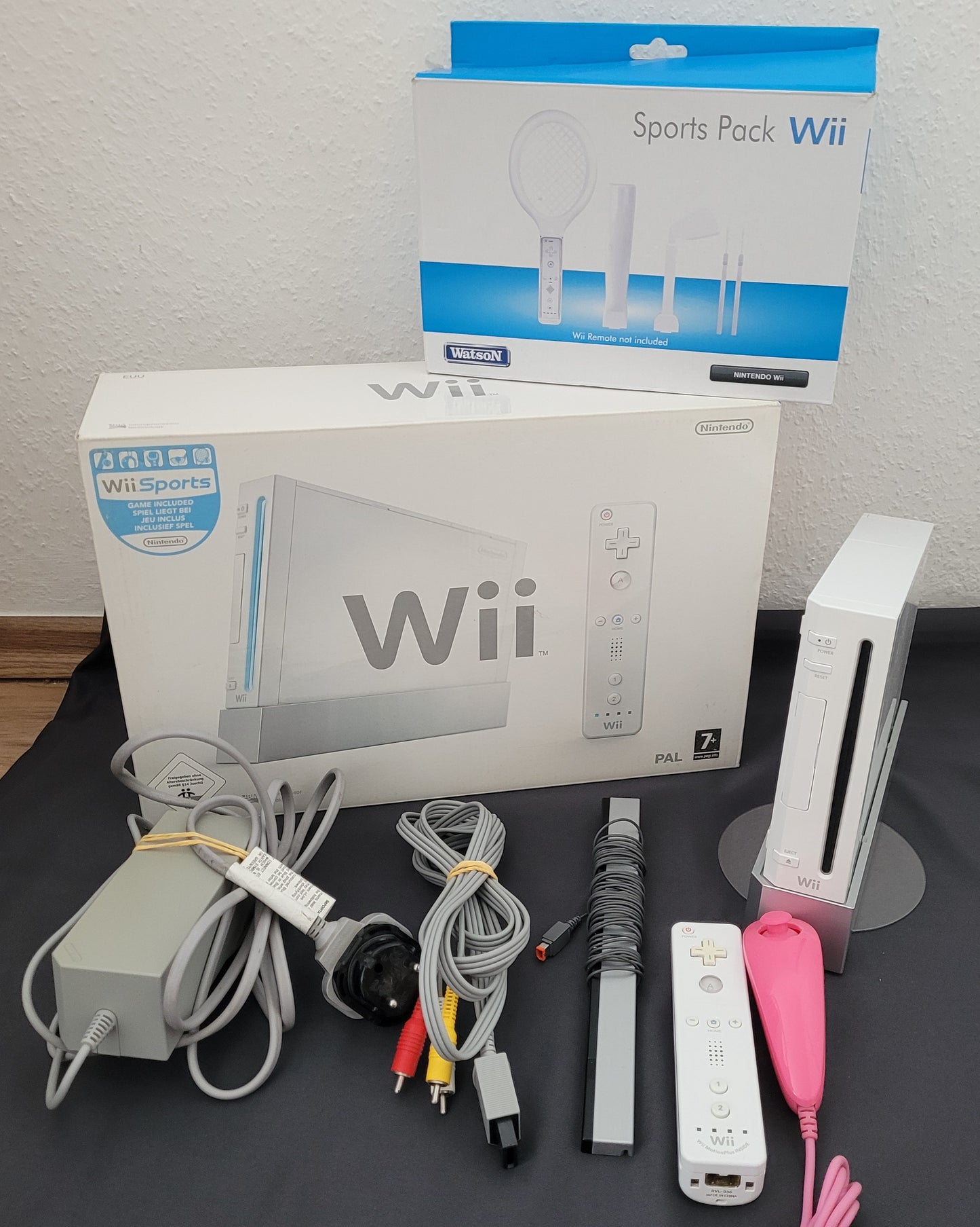 Die Vorderseite der Nintendo Wii Konsole Modell RVL-001 mit Zubehör sowie dem Wii Sports Pack. Die Konsole steht aufrecht auf einem Standfuß, begleitet von einem Netzteil, einem AV-Kabel, einer Sensorleiste und einer Wii-Fernbedienung mit pinker Schutzhülle. Daneben befindet sich die Verpackung des Wii Sports Pack mit verschiedenen Controller-Aufsätzen für Tennis, Golf und Baseball. Der Hintergrund ist weiß, während der Untergrund schwarz ist.